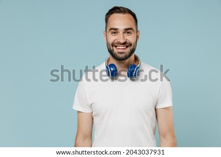 Young smiling happy satisfied man 20s wear blank print design white t-shirt headphones listen to music isolated on plain pastel light blue color background studio portrait. People lifestyle concept