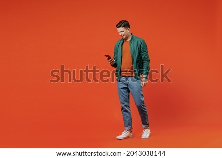 Full size body length smiling vivid happy young brunet man 20s wears red t-shirt green jacket hold in hand use mobile cell phone go step strolling isolated on plain orange background studio portrait.
