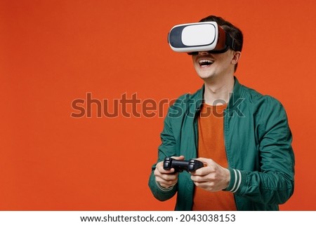 Smiling vivid excited happy young brunet man 20s wear red t-shirt green jacket watching in vr headset pc gadget play pc game with joystick console isolated on plain orange background studio portrait. Royalty-Free Stock Photo #2043038153