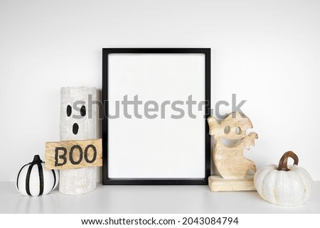 Halloween mock up. Black frame on a white shelf with rustic wood ghost decor and pumpkins. Portrait frame against a white wall. Copy space.