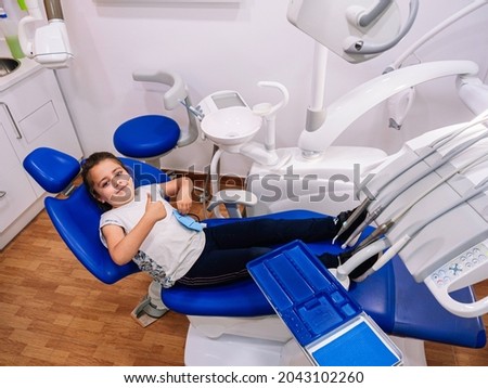 Little Caucasian girl is smiling, lying on the dental clinic chair and making the ok gesture with her hand.