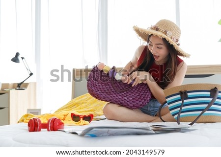 Asian young happy female traveler preparing belongings for sea and beach vacation trip with handbag, notebook, sunglasses, bikini, paper map and headphone on bed with excited and energetic happy face.