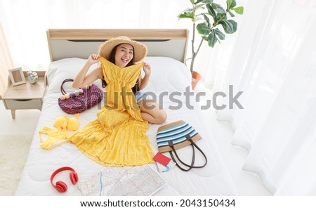 Top view shot of Asian young happy female traveler wears big hat laughing smiling look at camera sitting trying on yellow summer long dress on bed preparing personal stuff for dream destination trip.