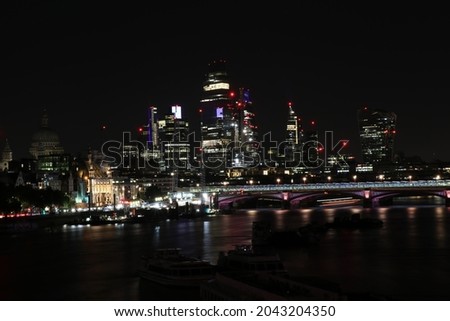 An urban skyline image of the City of London lit up at night, against a black sky.  Lights are reflected in the water of the River Thames.