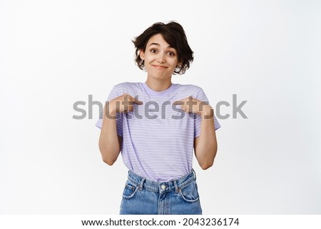 Image of cute modest girl pointing fingers at herself and smiling, its me gesture, self-promoting, talk about personal achievement, standing over white background