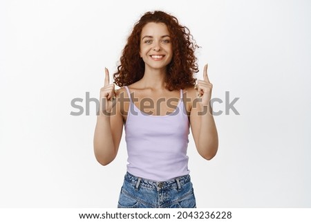 Portrait of smiling curly redhead woman, looking happy, pointing fingers up, showing advertisement, sale banner, standing over white background