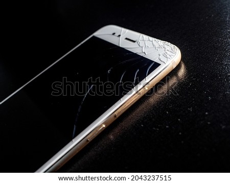 A thin white smartphone with a badly cracked screen, damage when falling. Close-up on a black background, the phone is on the table
