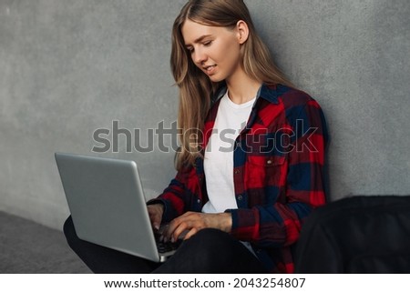 Happy young woman with backpack, in casual clothes, sitting on the floor with crossed legs and using laptop on gray background of city wall, outdoors