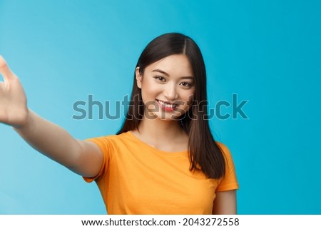 Close-up attractive friendly modern vietnamese girl extend hand, taking selfie, smiling joyfully, make photograph smartphone, hold camera arm, stand blue background joyful video-call friend Royalty-Free Stock Photo #2043272558