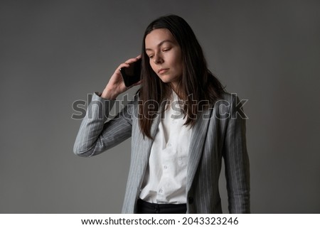 businesswoman dressed in a jacket and trousers in a casual style uses a smartphone for work and contact with partners, studio portrait on a gray, phone conversation, calmly listen to the interlocutor