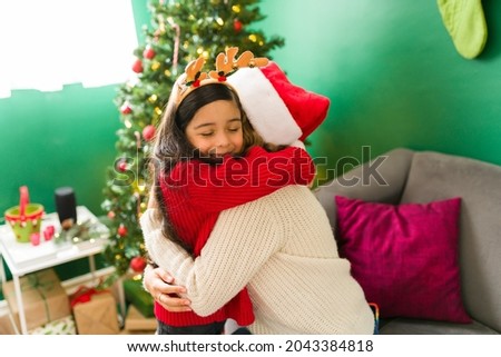 I love you mom. Adorable latin girl hugging her lovely mom with a santa hat next to the Christmas tree