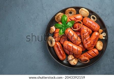 A plate of fried sausages with onions, garlic and basil on a dark gray background. Top view, space for copying. Royalty-Free Stock Photo #2043392159