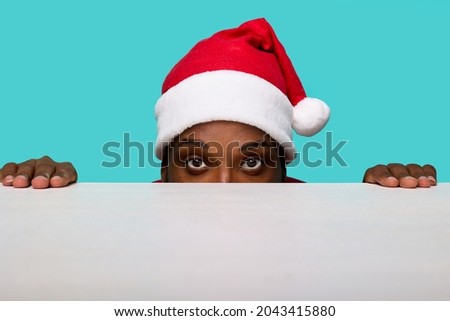 African-American man in a Santa Claus hat with expressive eyes looks intently at the camera looking out from under a white table on a turquoise background