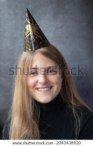 Close up of happy smiling teenager girl in a Halloween paper cap on a dark background. People, Halloween, decoration concept.