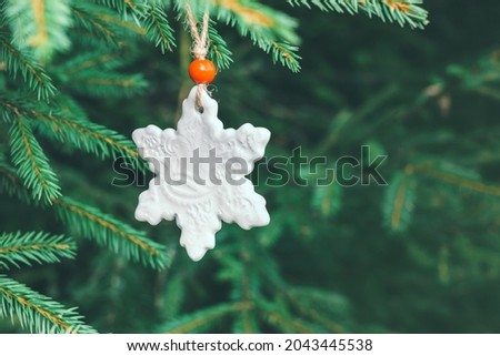 Unusual ceramic white toy in the shape of a snowflake with a red bead on a spruce branch. Minimalistic concept
