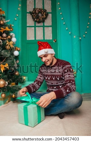 The guy in the burgundy sweater in the Scandinavian style in the New Year's interior. A man in a Santa hat unties a gift box. A surprise awaits