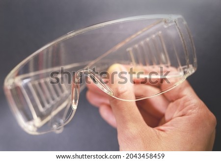 Protective glasses in hand, healthcare and safety concept. Safety glasses on black background. Personal protective equipment. Plastic goggles, transparent glasses safety at work Royalty-Free Stock Photo #2043458459