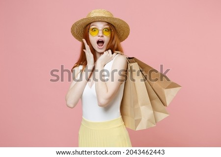 Young shocked surprised redhead ginger woman 20s wear straw hat glasses summer clothes holding package bags with purchases after shopping spread hand isolated on pastel pink background studio portrait