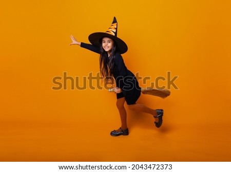 Little girl dressed as a Halloween witch in a black dress and hat flies on a broomstick on a yellow background.