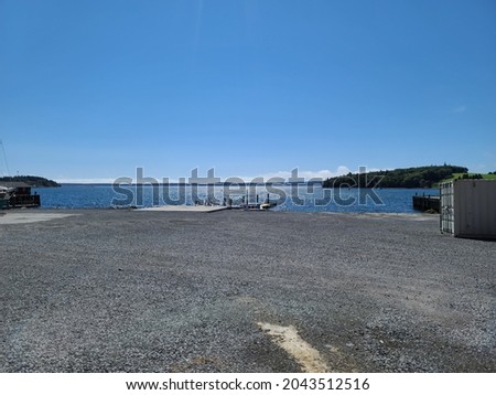A wide open gravel pit for parking near a wharf. The wooden dock extends into the calm water. Across the sea is dense, Nova Scotian forest made of evergreen pine trees.