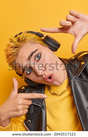 Surprised hipster girl searches perfect perspective makes hand frames measures angle has astonished expression vivid makeup yellow hair listens favorite music takes picture of moment poses indoor