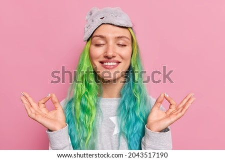 Peaceful smiling young woman with dyed colorful hair keeps eyes closed makes mudra gesture relaxes after studying or work meditates before sleep wears nightwear poses against pink background