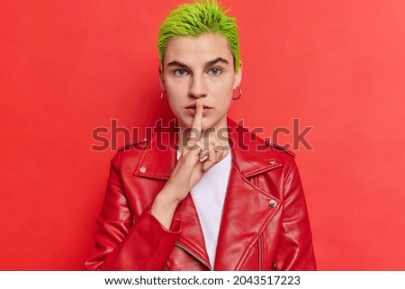 Keep silence please. Serious hipster girl with green hairstyle makes shush gesture keeps index finger over lips zips wears leather jacket poses against vivid red background. Hush its my secret Royalty-Free Stock Photo #2043517223