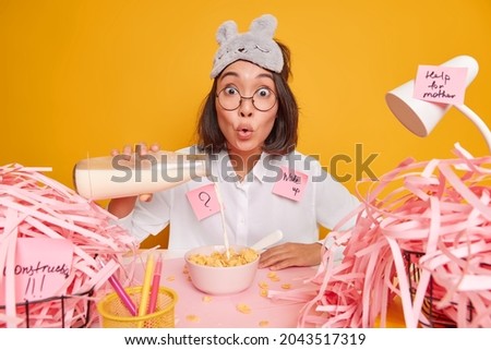 Photo of surprised young Asian woman pours milk in cornflakes prepares breakfast makes memo stickers has mess on desktop isolated over yellow background has busy schedule. Office work concept