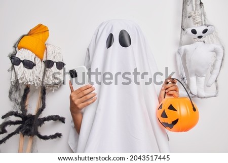 Unrecognizable female ghost holds modern mobile phone and carved pumpkin prepates for Halloween celebration searches in internet ideas to decorate room before party poses near scary toys indoor.
