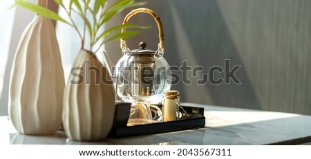 glass tea pot on wooden tea tray with white flower vase on marble dining table with asun shade in living room home background concept