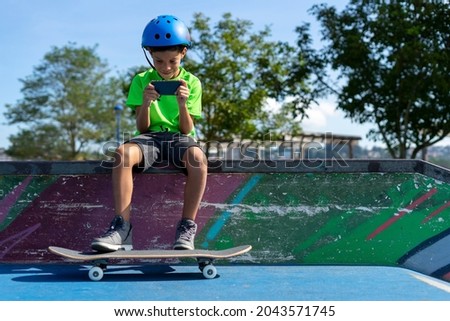 Boy watches videos of skateboarders on cell phone while taking a break after practicing on his skateboard in the park. . High quality photo