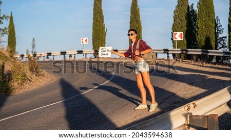 Woman try to stop car with cardboard sign, copy space. Girl with cheerful face travelling by hitchhiking with road on background. Travelling and hitchhiking concept.