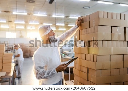 The food factory inspector in a white sterile uniform taking boxes on from a pile. He is holding a tablet. If the content of the box is good, the whole pile is ready for shipping. Quality control