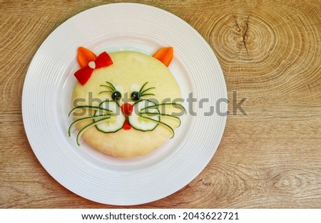 Cute cat face made it from bread,cucumbers,tomatoes,carrots,black olives and white sesame on plate with wooden background.Art food idea for kids breakfast.Top view.Copy space