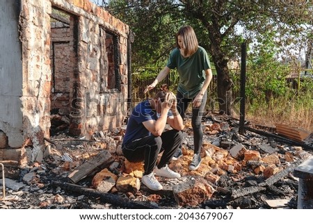 Young couple on the ruins of house destroyed with fire. Damaged window, walls and floor, burnt wooden frame. Man sits wrapping head in hands, woman calms him. Insurance or emergency concept. Royalty-Free Stock Photo #2043679067