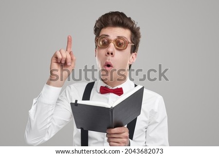 Funny young male genius in elegant outfit with bow tie and nerdy spectacles, holding open notebook and pointing finger up while having brilliant idea against gray background