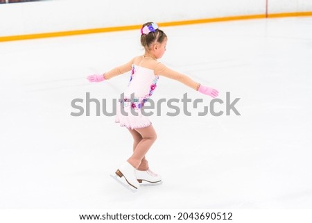 Little girl in a pretty pink dress with flowers practicing figure skating moves on an indoor ice rink.