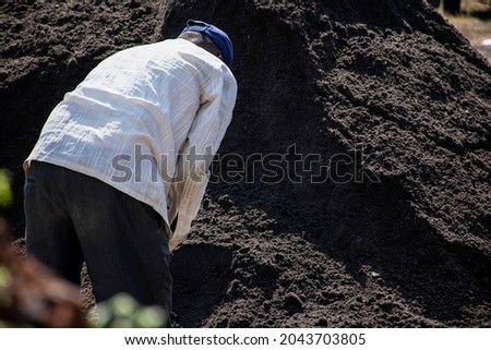Stock photo of 50 to 60 year old Indian construction worker shoveling the black sand or black soil with a spade under bright sunlight at Kolhapur, Maharashtra, India.
