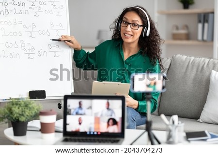Videoconference And Training. Class of diverse students learning remotely online using web app. Female teacher in headset providing lesson looking at pc screen, recording video on cellphone on tripod Royalty-Free Stock Photo #2043704273