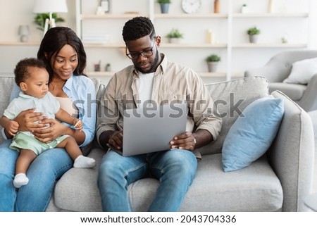 Portrait of African American baby boy sitting with parents on sofa and looking on laptop, watching cartoons. Video call with grandparents. Dad showing his son pictures on computer screen