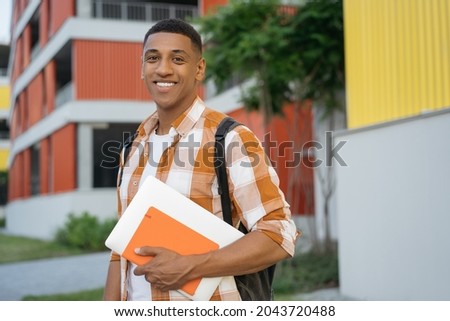 Authentic portrait of confident developer holding book and laptop walking on urban street. Handsome smiling African American student student looking at camera in university campus. Education concept