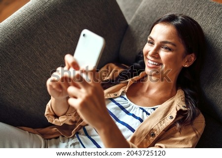 A woman lies on the couch with a phone in the living room. Communication, online shopping. A woman with a smartphone communicates, rests and has fun. Royalty-Free Stock Photo #2043725120