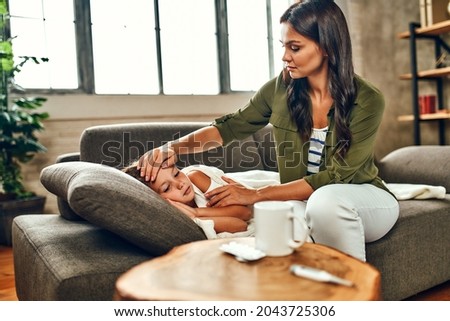 The child has a high fever. Mom touches the forehead of her daughter, who is lying under the covers on the couch and is sick. Royalty-Free Stock Photo #2043725306