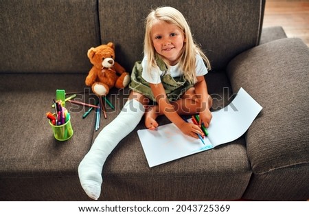 A cute little girl with a broken leg in a cast is sitting on the couch at home and is drawing.