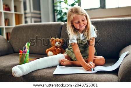 A cute little girl with a broken leg in a cast is sitting on the couch at home and is drawing.