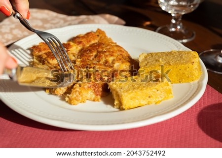 Hand of a caucasian woman eating frico, typical eastern Italy dish with mixed cheeses and golden cornmeal mush on a white plate under the sunlight at lunch time Royalty-Free Stock Photo #2043752492