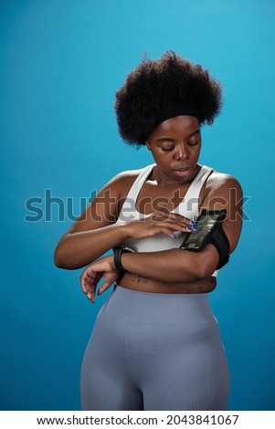 Young sportswoman in activewear touching screen of smartphone on her arm against blue background