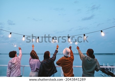 Back view of young ecstatic friends with bengal lights enjoying summer evening by lake Royalty-Free Stock Photo #2043841289