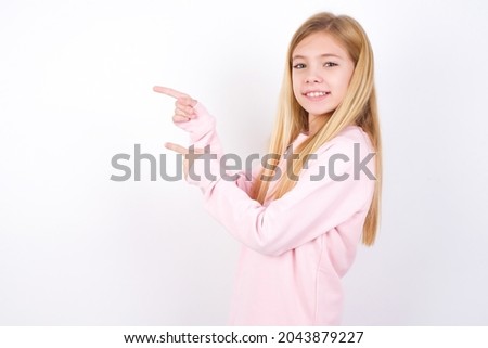 beautiful caucasian little girl wearing pink sweater over white background points aside with  surprised expression with mouth opened, shows something amazing. Advertisement concept.