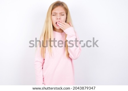 Sleepy beautiful caucasian little girl wearing pink sweater over white background yawning with messy hair, feeling tired after sleepless night, yawning, covering mouth with palm.